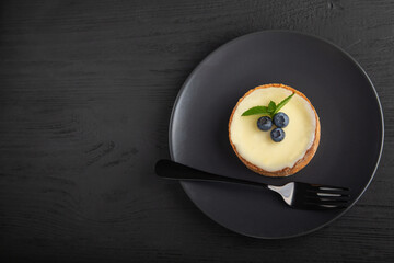 Tasty cake or tartlet with berries, vanilla cream and mint on a black plate with fork, copy space on black wooden background, top view