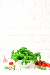 Fresh basil in a bowl and red tomatoes. Kitchen garden on a white table, brick wall. Copy space for text
