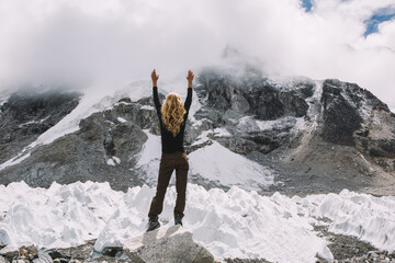 Young female tourist enjoying beautiful view in foggy Himalayas.Back view of woman holding hands up while climbing in high mountains glacier.Traveller standing on edge of beautiful canyon