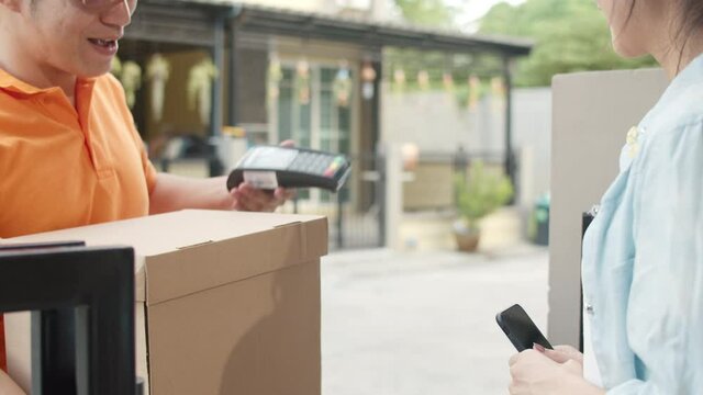Young postal delivery courier man holding parcel boxes for sending to customer, Asian women signing and pay via qr code on mobile phone for receive delivered parcels outdoors. 4k Slow motion.