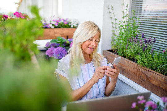 Active senior woman with laptop and smartphone working on balcony, home office.
