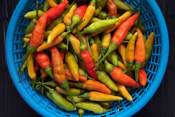 Top view closeup Heap of various colors fresh raw Thai chili peppers in a plastic blue basket with black bamboo placemat background.
