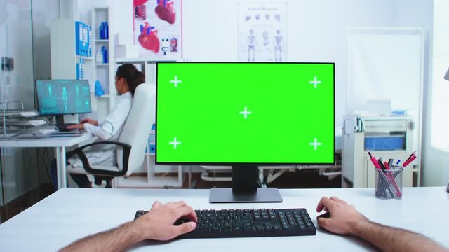 Pov of doctor hand working on computer with green screen in hospital cabinet. Medical physician in clinic cabinet working desktop with copy space available.
