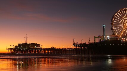 Twilight waves against classic illuminated ferris wheel, amusement park on pier in Santa Monica pacific ocean beach resort. Summertime iconic symbol of California glowing in dusk, Los Angeles, CA USA.