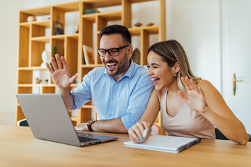 Smiling couple waving at web camera using laptop for video call.