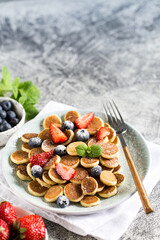 Tiny pancakes for breakfast. Cereal pancakes with blueberries, strawberries in plate on grey background. Trendy food. Copy space for text or design