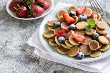 Tiny pancakes for breakfast. Cereal pancakes with blueberries, strawberries in plate on grey background. Trendy food. Copy space for text or design