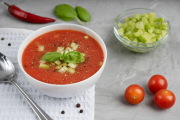 Red gazpacho soup made of pureed tomatoes decorated with basil leaves and cucumber in a bowl standing on white towel on concrete surface surrounded by peppercorns and spoon. Horizontal orientation