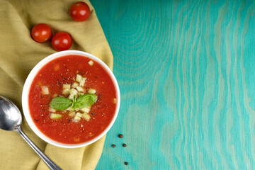 Gazpacho soup made of red ripe pureed tomatoes decorated with basil leaves and cucumber in white bowl standing on olive towel on blue wooden table together with silver spoon. Image with copy space