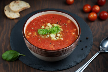 Red gazpacho soup made of pureed tomatoes and other vegetables decorated with green basil leaves and copped cucumber in a bowl standing on stony tray on dark brown wooden table at kitchen