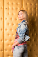 A blonde girl in a denim suit stands half-turned and poses against the background of a golden wall.