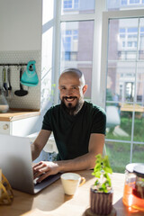 The guy works from home. He sits in the kitchen at the table and uses a laptop computer and drink hot tea. He is smiling, his business is going well.