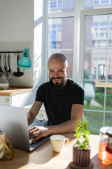 Freelancer programmer working from home on kitchen and using laptop. Bearded man working with a computer, prints text on keyboard. Self entrepreneur sitting and working at his modern apartment.