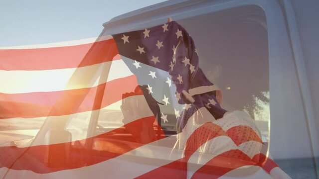 Animation of American flag waving over mixed race woman in car taking photos by seaside