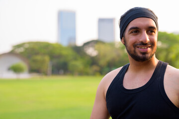 Face of happy young handsome bearded Indian man thinking at the park