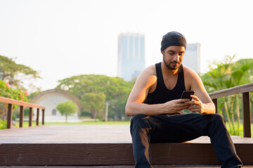 Young handsome bearded Indian man using phone while sitting at the park