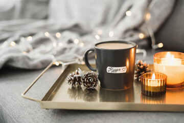 Hygge concept: black mug with hot coffee served on a metal tray, burning candles, pine cones as a...