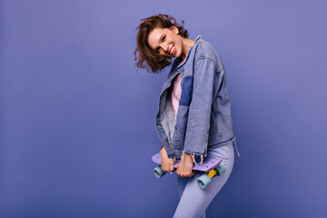 Charming woman with trendy haircut enjoying photoshoot with skateboard. Lovely girl with happy face expression laughing on violet background.