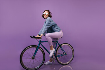 Magnificent female bicyclist posing in denim attire. Laughing debonair girl sitting on bicycle.