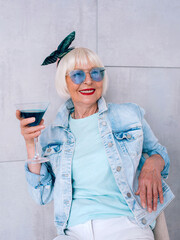 senior (old) stylish woman with gray hair and in blue glasses and denim jacket holding glass with...