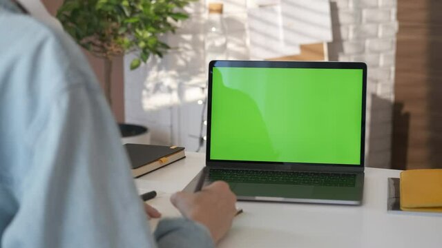 Young student looking at green screen laptop computer in living room watching education video content online. Over shoulder close up view. Business woman or female freelancer working from home office.