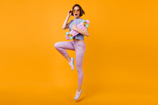 Full-length portrait of stunning girl in yellow-colored glasses expressing amazement. Indoor shot of cheerful brunette lady with longboard jumping on orange background.