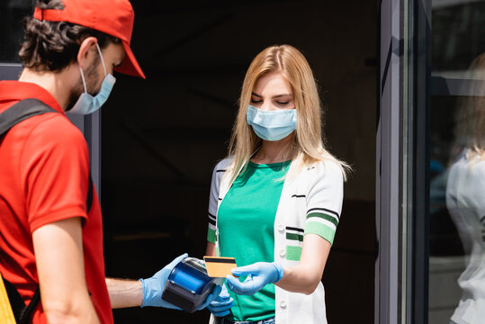 Selective focus of woman in medical mask paying with credit card near courier holding payment terminal on urban street