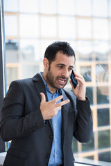 manager moro vestito con giacca nera e camicia blu, in piedi di fronte a una vetrata si arrabbia...
