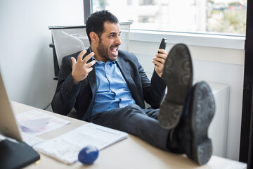 manager moro vestito con giacca nera e camicia blu, seduto nella sua postazione di lavoro tiene i piedi sulla scrivania e si stressa dopo una telefonata.