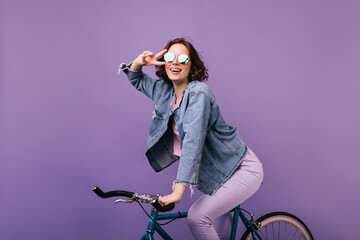 Pleasant stylish woman posing on purple background on bike. Indoor photo of smiling ecstatic girl sitting on bicycle.