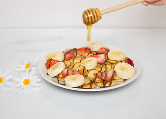 Trendy food - pancake cereal. Heap of mini cereal pancakes on a white plate on a white background. Honey drips onto the dish with a wooden stick, strawberries and bananas.