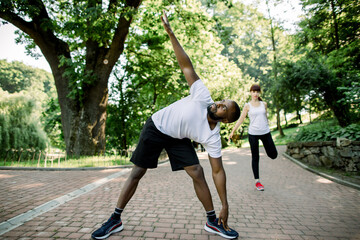 Obraz premium Sport, fitness and jogging concept. Young multietnic couple ready to run on fresh morning in the city park, stratching before exercises. Focus on African man with arm raised. Woman on the background