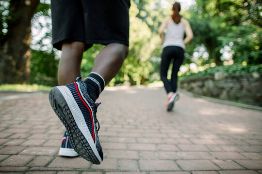 Close up back view photo of legs of African man in sport sneakers jogging in the city park on a sunny summer day, together with his female Caucasian friend, running on the background