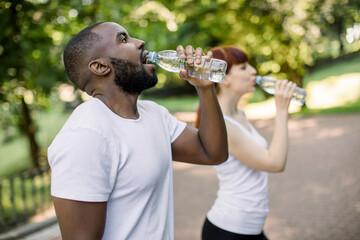 Multiethnical male and female friends drinking water from bottle after fitness sport exercise at city park. Smiling couple with bottles of cold drink outdoors. Focus on African man