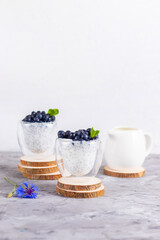 Obraz na płótnie Canvas Chia pudding with mint and blueberries in glasses on a wooden stand with blue chicory flower and white cream jug on a white background