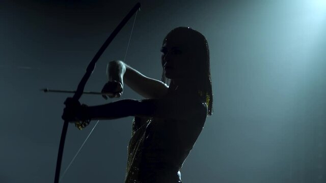 Silhouette of Joan of Arc with bow and arrow on stage in a dark studio with smoke and neon lighting. A bright young woman in a gold dress and a headdress. Dynamic neon lighting effects. Luxurious