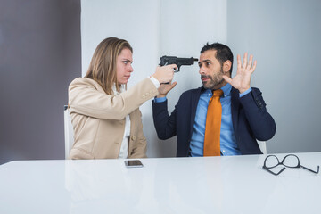 una donna bionda con giacca beige punta la pistola al suo collega che reagisce terrorizzo alzando...