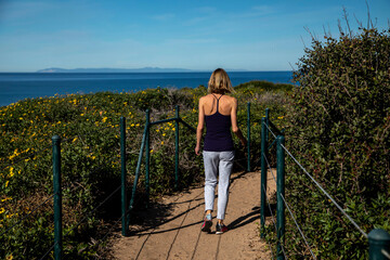 Short haired blonde women in her 40s in jeans and black top walking on dirt trail overlooking the pacific ocean flanked by daisy bushes