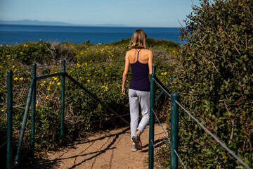 Short haired blonde women in her 40s in jeans and black top walking on dirt trail overlooking the pacific ocean flanked by daisy bushes