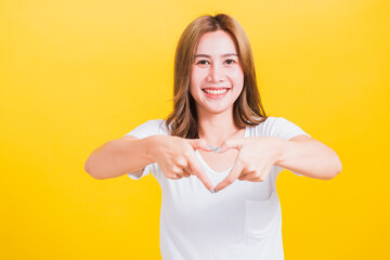 Fototapeta na wymiar Asian Thai happy portrait beautiful cute young woman smile standing make finger heart figure symbol shape sign with two hands and looking camera, studio shot isolated yellow background with copy space