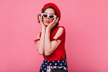 Attractive french girl with pale skin posing in studio. Indoor photo of lovable young lady in sunglasses standing on rosy background.