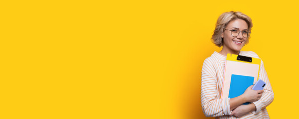 Charming blonde student with eyeglasses holding some folders smiling at camera on a yellow wall...