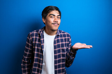 Portrait of young Asian student wearing shirt smiling and pointing to presenting something on his side, against blue background with copy space