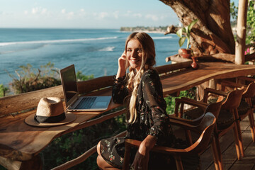Young business woman working at the computer in cafe wit sea view, look at the camera and smiling. Technology and travel