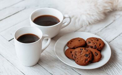 Breakfast in the morning. Two white cups of coffee and chocolate chip cookies stand on a light wooden parquet and pillows lie nearby. Homeliness, day off. Pastel colors.