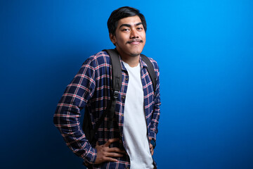 Asian students wearing backpack smiled at the camera looking confident while putting both hands on the waist against blue background. The student is ready to go to school or college. Studio shots