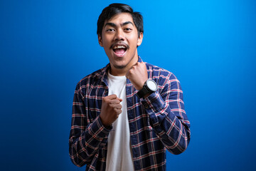 Naklejka premium Joyful young man student in casual clothes isolated on blue background studio raised his arm to celebrate his victory. The guy dance happily over the good news he received.