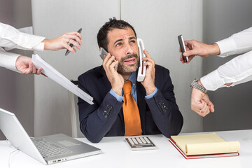Manager i giacca e cravatta seduto nella postazione di lavoro del suo ufficio è circondato da mani e impegni e reagisce in maniera stressata mentre risponde al telefono - obrazy, fototapety, plakaty
