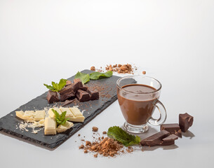 Chunks of dark and white chocolate presented on a slate board with mint leaves. Cup of chocolate on a white background.