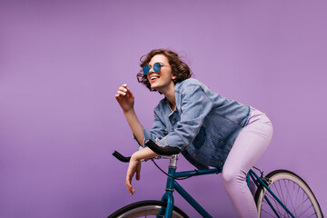 Carefree short-haired lady sitting on bicycle in studio. Happy caucasian girl with wavy hairstyle expressing positive emotions.